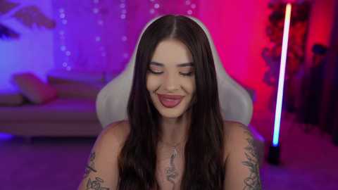 Media: Video of a smiling, topless woman with long dark hair, tattoos, and wearing a white veil, in a dimly lit room with purple and red lighting, leaning on a white chair.