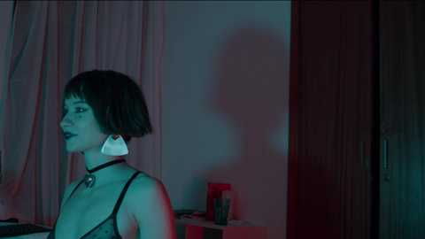 Media: Video of a pale-skinned woman with short black hair, wearing a black choker and a large, white, geometrically shaped earring, standing in a dimly lit room with red and blue lighting, reflecting off a wall.