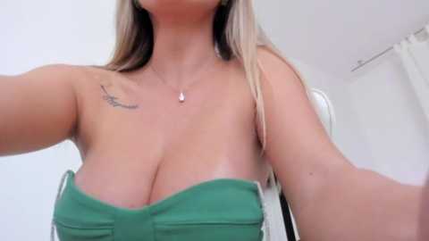 Media: A video of a light-skinned woman with long, straight blonde hair, wearing a strapless green top that reveals her ample cleavage. She has a small bird tattoo on her right shoulder. The background is a plain white room with a white curtain.