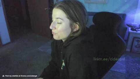 Media: A video of a young woman with light skin and shoulder-length brown hair, eyes closed in meditation, seated in a dark room with a wooden floor and a computer monitor on the right.