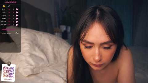 Media: Video of a young woman with long black hair and fair skin, wearing minimal makeup, lying on a bed with white sheets. The background features a dimly lit room with a nightstand and a potted plant.