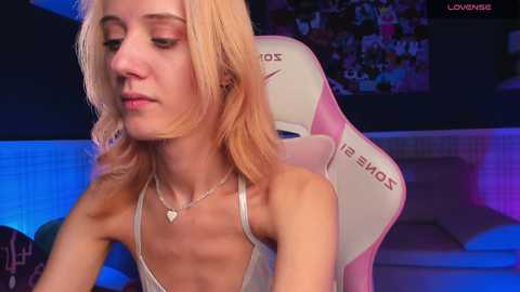 Media: Video of a slender, fair-skinned, blonde woman with a heart-shaped necklace, wearing a thin, revealing tank top, seated in a pink gaming chair in a dimly lit room.