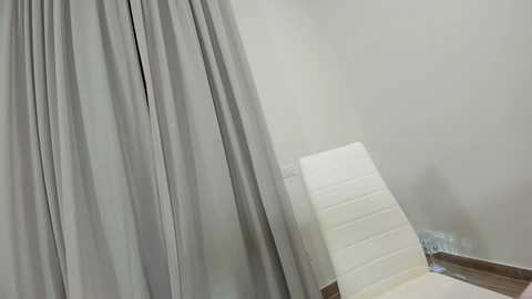 Media: A video of a minimalist, modern room featuring a gray curtain and a white massage table against a plain white wall, with a wooden floor and a power outlet visible.