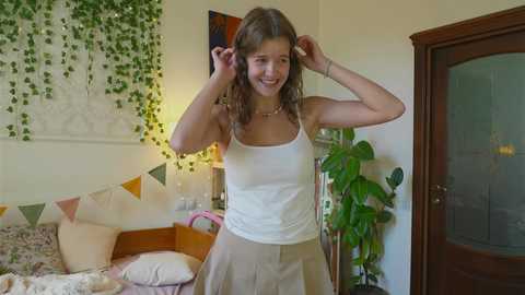 Media: Video of a smiling young woman with light skin and wavy brown hair adjusting her headband, wearing a white tank top and beige skirt, standing in a cozy bedroom with green ivy wall art, wooden door, and potted plants.