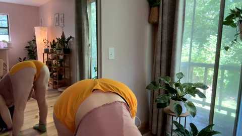 Media: Video of a woman in a bright yellow sports bra and pink shorts bending over in a living room with large windows, indoor plants, and a wooden floor.