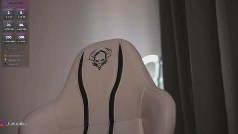 Media: Video of a white gaming chair with a black skull logo and black accents, in a dimly lit room with muted gray curtains.