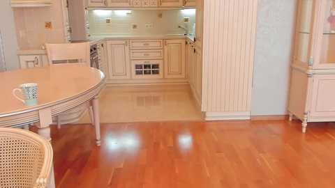 Media: A video of a quaint, vintage-style kitchen with light wood cabinets, a white door, and a polished wooden floor. A round table with a light blue cup and a wicker chair are visible in the foreground.