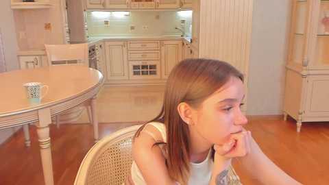 Media: Video of a young, light-skinned girl with straight brown hair, wearing a white tank top, sitting on a woven chair, looking contemplative in a cream-colored kitchen with light wood cabinets and a round table.