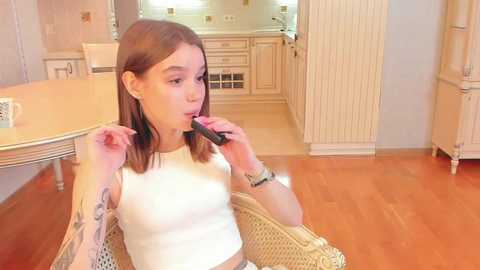 Media: A video of a young Caucasian woman with shoulder-length brown hair, wearing a white crop top, sitting on a wicker chair and applying lipstick. The background features a light-colored kitchen with wooden flooring.