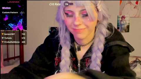 Media: Video of a pale-skinned woman with long, platinum-blonde braided hair, wearing a black hoodie, and pink makeup, smiling at the camera in a dimly lit room.