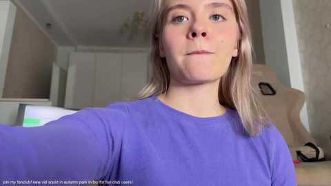 Media: A video of a young woman with light skin and straight, shoulder-length blonde hair, wearing a purple long-sleeved shirt, in a modern, well-lit room with beige walls, a white door, and a gaming chair.