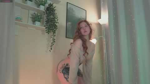 Media: Video of a curvaceous, fair-skinned woman with long, curly red hair, wearing a sheer, off-the-shoulder top, leaning against a white wall adorned with potted plants.