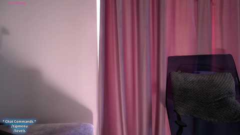 Media: Video of a modern room with soft pink and white curtains, a grey armchair with a textured pillow, and a shadowy corner.