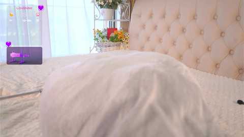 Media: Video of a plush, white bed with tufted, pastel pink headboard in a bright, airy room with sheer white curtains and a floral arrangement on a shelf.