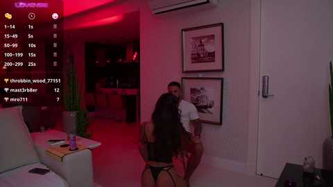 Media: Video of a dimly-lit modern bedroom with red lighting, featuring a man in a black robe hugging a woman in lingerie against a wall adorned with framed photos.