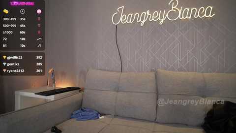 Media: Video of a dimly lit, modern living room featuring a beige couch, a white side table, a glowing \"Joangrey Bianca\" neon sign, and a discarded blue shirt on the floor.