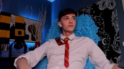 Media: Video of a young, slim, fair-skinned man with short brown hair, wearing a white shirt and red striped tie, sitting on a blue fluffy chair in a modern, eclectic room with abstract art and a black and white butterfly wall tapestry.