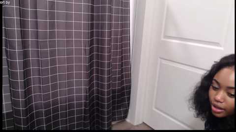 Media: Video of a young Black woman with curly hair peering around a closed white door, gazing at a black-and-white checkered shower curtain.