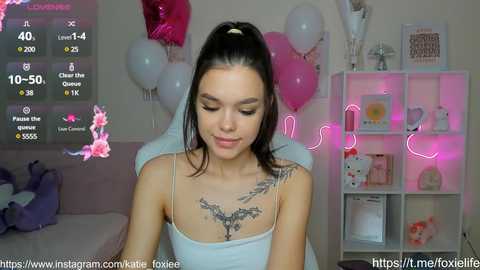 Media: Video of a young woman with fair skin, dark hair, and a tattoo on her chest, seated in a pastel-themed room with pink balloons, neon lights, and shelves filled with toys and decor.