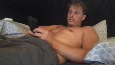 Media: Video of a shirtless, muscular man with short blond hair, lying on a bed with gray and white checkered sheets, holding a remote control, in a dimly lit room with black curtains.