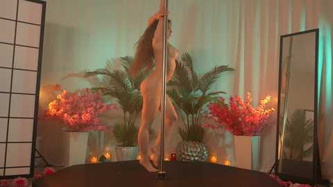 Media: Video of a nude woman dancing sensually on a pole, surrounded by pink flowers and lit candles in a dimly lit room with a mirrored background.