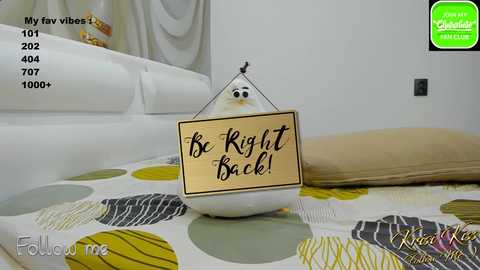 Media: Video of a plush duck with a sign saying \"Be Right Back!\" on a bed with a white sheet and yellow, green, and gray polka dots.