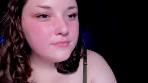 Media: Video of a young woman with fair skin, curly brown hair, wearing a green top, against a dark background. Her expression is neutral, and her cheeks have a slight blush.