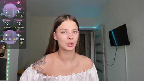 Media: Video of a young woman with long brown hair and fair skin, wearing a white off-shoulder top, standing in a modern room with a TV on the wall.