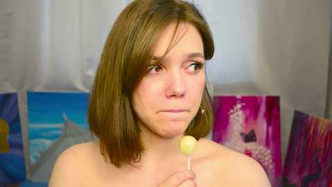 Media: Video of a young woman with shoulder-length brown hair, holding a yellow lollipop, with a melancholic expression. Background features colorful, abstract paintings.