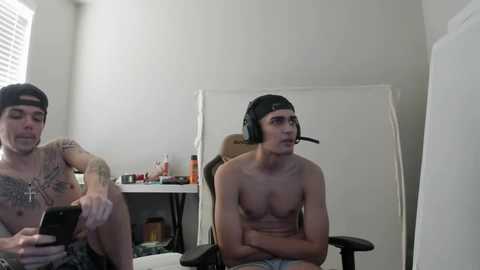 Media: Video of two shirtless men with tattoos, one wearing a backward cap and headphones, sitting in a dimly lit room with a whiteboard and cluttered desk.