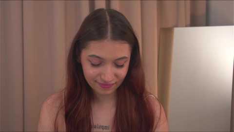 Media: Video of a young woman with long, straight brown hair, fair skin, and a delicate, slender frame. She wears a light-colored top and looks down, appearing introspective. Background features beige curtains and a white lamp.