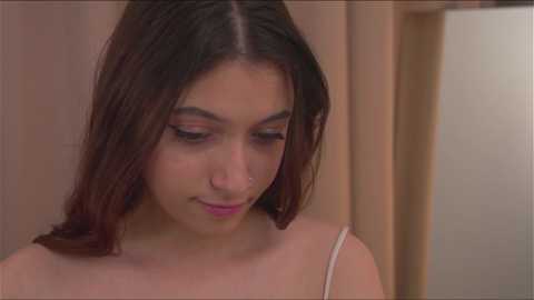 Media: Video of a young Latina woman with straight, shoulder-length dark hair, wearing a light-colored spaghetti-strap top. She has a nose stud and is looking down, appearing contemplative. The background is blurred with beige and light brown tones.