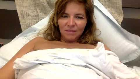 Media: Video of a middle-aged woman with light skin and shoulder-length brown hair, lying in a hospital bed covered in white sheets, with a beige cloth backdrop. She has a neutral expression.