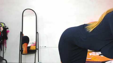 Media: Video of a woman with long blonde hair bending over, facing a tall mirror with a black frame, in a minimalist room with white walls and orange objects on the floor.