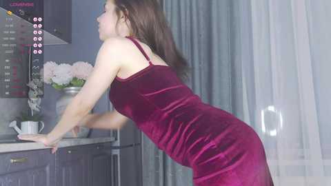 Media: Video of a slender, light-skinned woman with long brown hair, wearing a deep red velvet dress, leaning over a kitchen counter, looking at a stove, with gray and white curtains in the background.