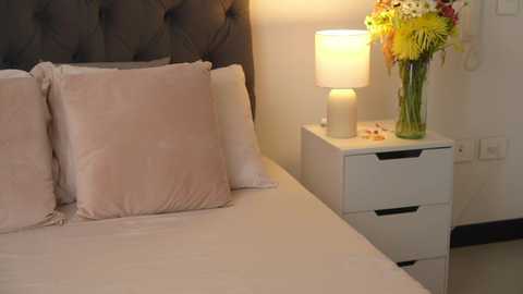 Media: Video of a cozy bedroom with a plush, light pink bed, gray upholstered headboard, and a nightstand holding a vase of colorful flowers.