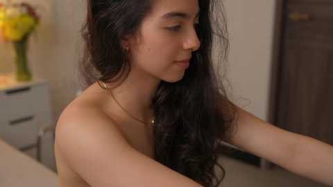 Media: Video of a young woman with long, wavy black hair, wearing a thin gold necklace, standing topless in a modern kitchen with beige walls and a white countertop, holding a glass of water.