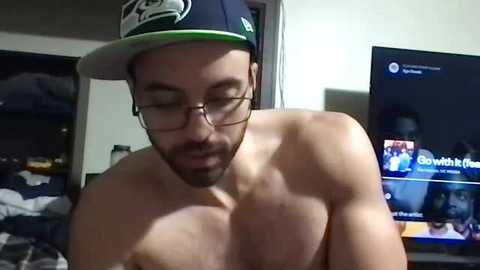 Media: Video of a shirtless, muscular white man with glasses, wearing a dark baseball cap, eyes closed, sitting in a dimly lit room with a TV showing a social media feed in the background.