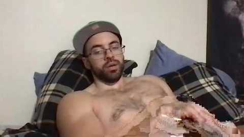 Media: Video of a shirtless, bearded man with glasses and a cap, lying on a bed with plaid pillows, holding a glass, against a plain wall.