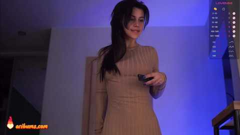 Media: A video of a slender, fair-skinned woman with long, dark hair, wearing a beige, ribbed long-sleeve dress, holding a remote, standing in a dimly lit room with blue lighting and a modern staircase.