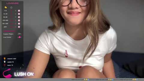 Media: Video of a young woman with long, straight, light brown hair, wearing glasses, a white T-shirt, and shorts, smiling in a dimly lit room.