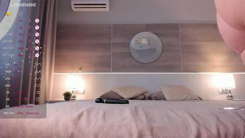 Media: Video of a modern bedroom with a minimalist design: light wood-paneled walls, a round mirror, two bedside lamps, an air conditioner, and a digital clock on the left.