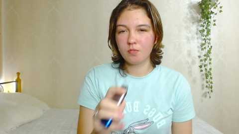 Media: Video of a young woman with shoulder-length brown hair, wearing a light blue t-shirt, blowing on a silver mobile phone in a beige room with a bed, green plant, and a lamp.