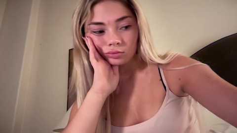 Media: A video of a young Asian woman with long blonde hair, wearing a white tank top, resting her head on her hand, in a bedroom with a bed and beige wall.