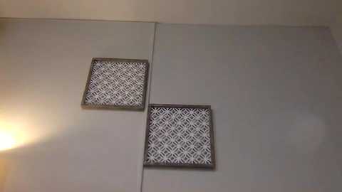 Media: Video of two decorative wall panels with geometric white patterns on beige backgrounds, mounted side by side on a smooth, light beige wall. The left panel is slightly lower than the right, creating a slight diagonal line.