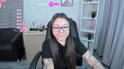 Media: Video of a smiling, tattooed woman with long dark hair and glasses, wearing a black top, seated in a black office chair in a light-colored room.