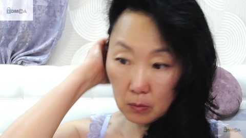 Media: Video of an Asian woman with long black hair, wearing a light purple bra, resting her head on a pillow, with a white and patterned pillow in the background.