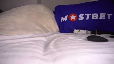 Media: A video of a bed with a white pillow and a blue pillow with \"MOSTBET\" logo. A black remote control and a white and black container are on the bed.