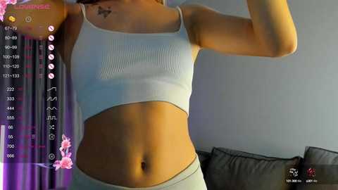 Media: Video of a slim, light-skinned woman wearing a white sports bra and matching high-waisted shorts. She has a small butterfly tattoo on her left collarbone. Background includes a gray couch and a pink floral filter overlay.