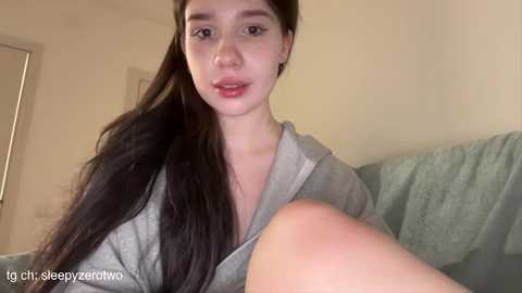 Media: Video of a young woman with long, dark hair, fair skin, and red lipstick, wearing a gray hoodie, sitting on a light blue couch in a dimly lit room.